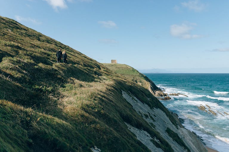 Serene coastal scene with two people walking along a grassy cliffside path under a clear blue sky.